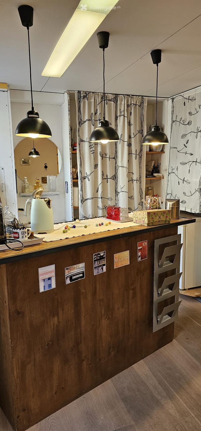 Modern kitchen with wooden countertop, pendant lights, and various kitchen items including a teapot, cups, and a refrigerator.
