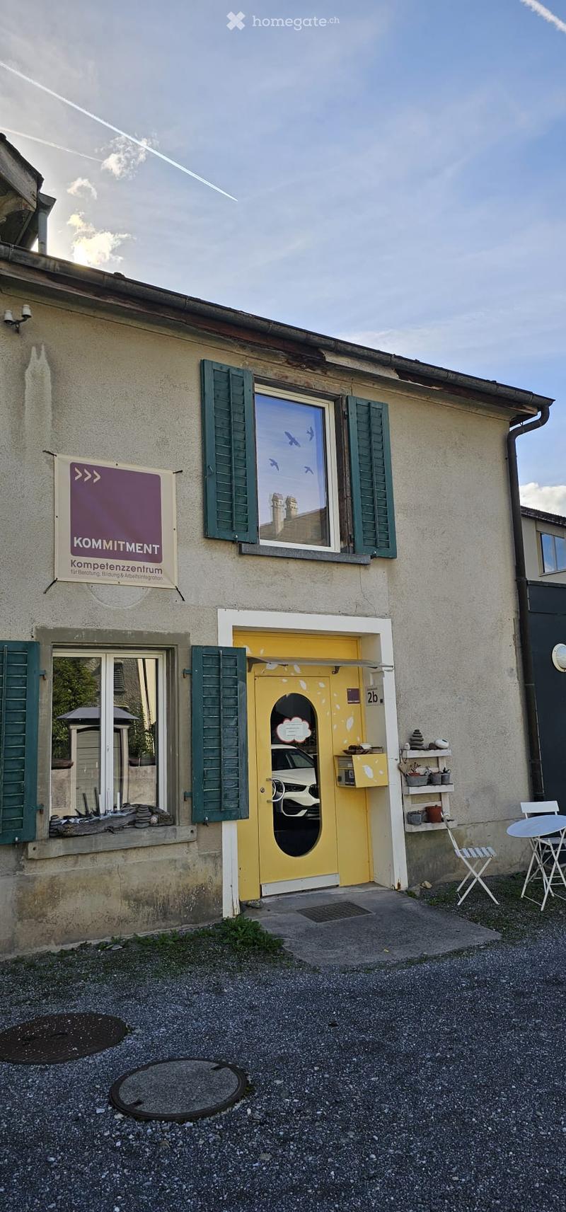 Two-story building with a yellow door, green shutters, and a sign reading "Kompetenzzentrum" on the wall. A small outdoor seating area with a table and chairs is visible to the right.