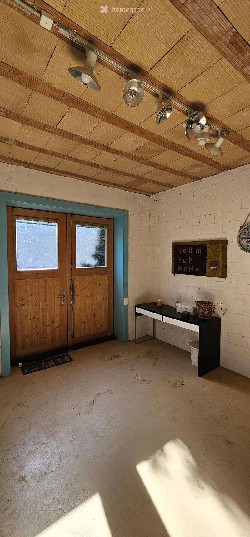 A small, unfurnished room with a wooden door, a table with a basket, and a clock on the wall.