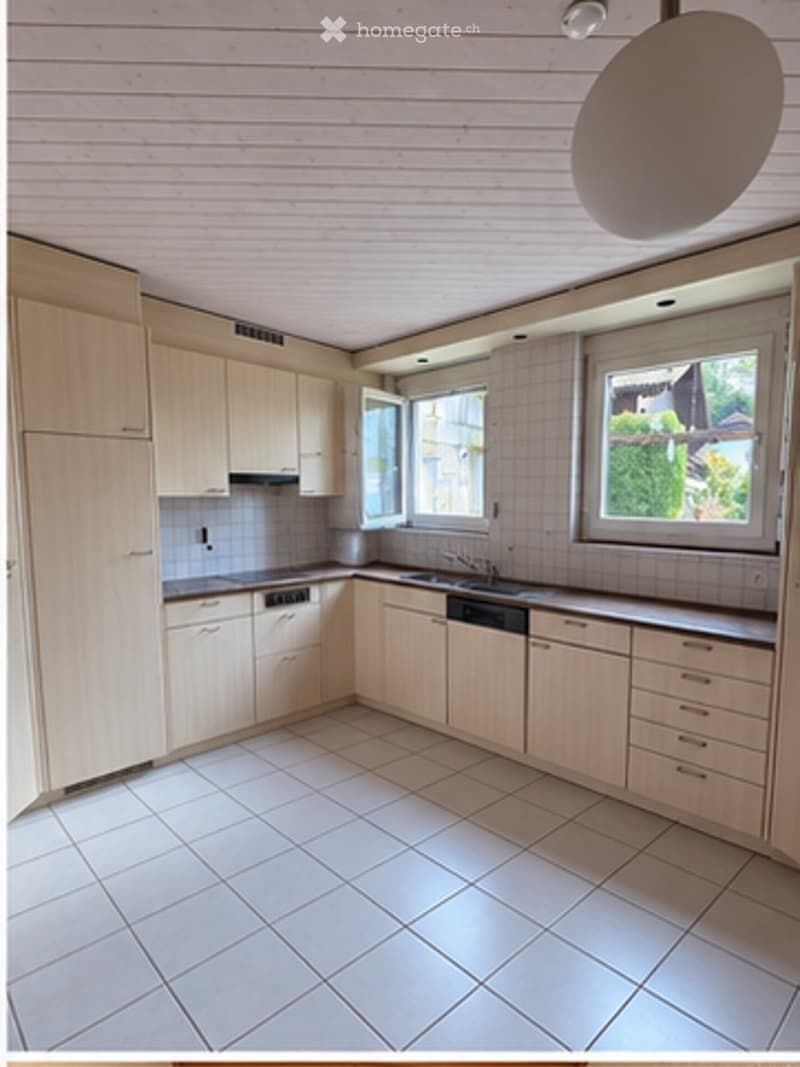 Bright, modern kitchen with light wooden cabinets, tiled floor, and stainless steel appliances including a dishwasher. Large windows provide natural light.