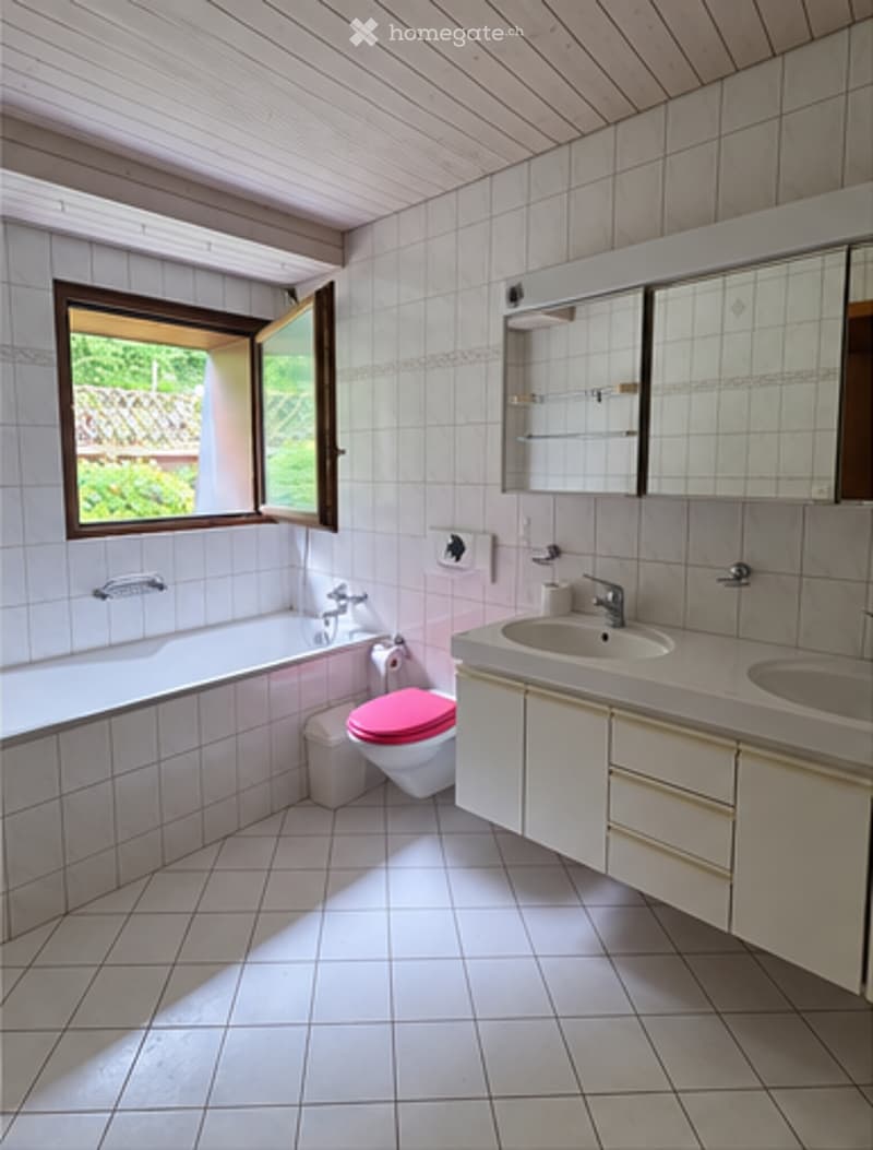 Bright bathroom with white tiled walls and floor, a bathtub, a toilet with a pink seat cover, a double sink with a mirror above, and a window letting in natural light.
