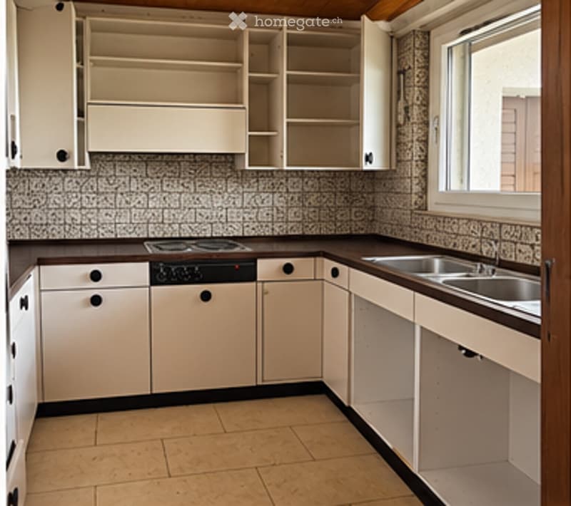 A clean, unfurnished kitchen with white cabinets, a stainless steel sink, and a stove. The space is well-lit with natural light from a window on the right side.