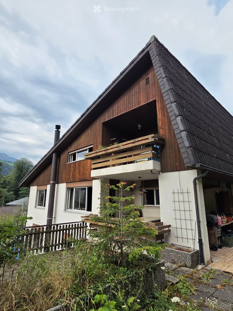 Two-story house with wooden exterior, balcony, and garden area with plants and a small table.