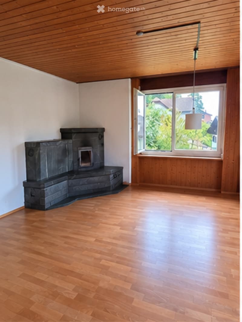 Empty room with wooden flooring, white walls, and a fireplace. Large window with a view of greenery outside.