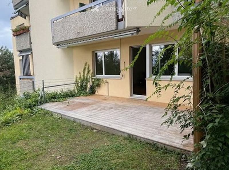 Two-story building with a balcony, wooden deck, and greenery around the entrance.