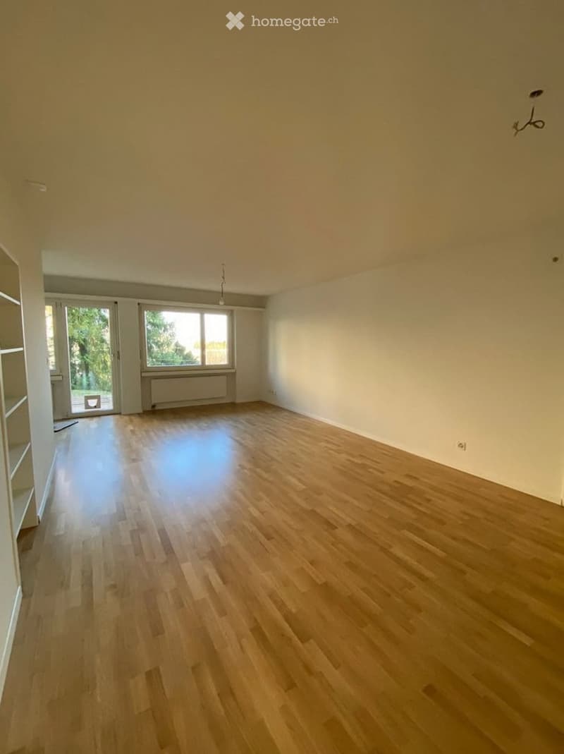 Empty room with wooden flooring, white walls, large windows, and a sliding glass door leading to an outdoor area.