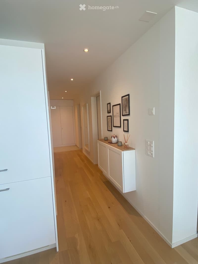 A modern hallway with light wooden flooring, white walls, and recessed lighting. Features a small white console table with decorative items and framed artwork on the wall.