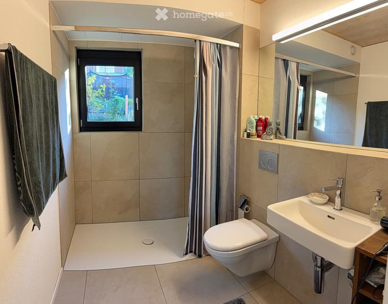 Modern bathroom with a walk-in shower, toilet, sink, and large mirror. Beige tiles, a window with a view of greenery, and a towel hanging on the wall.