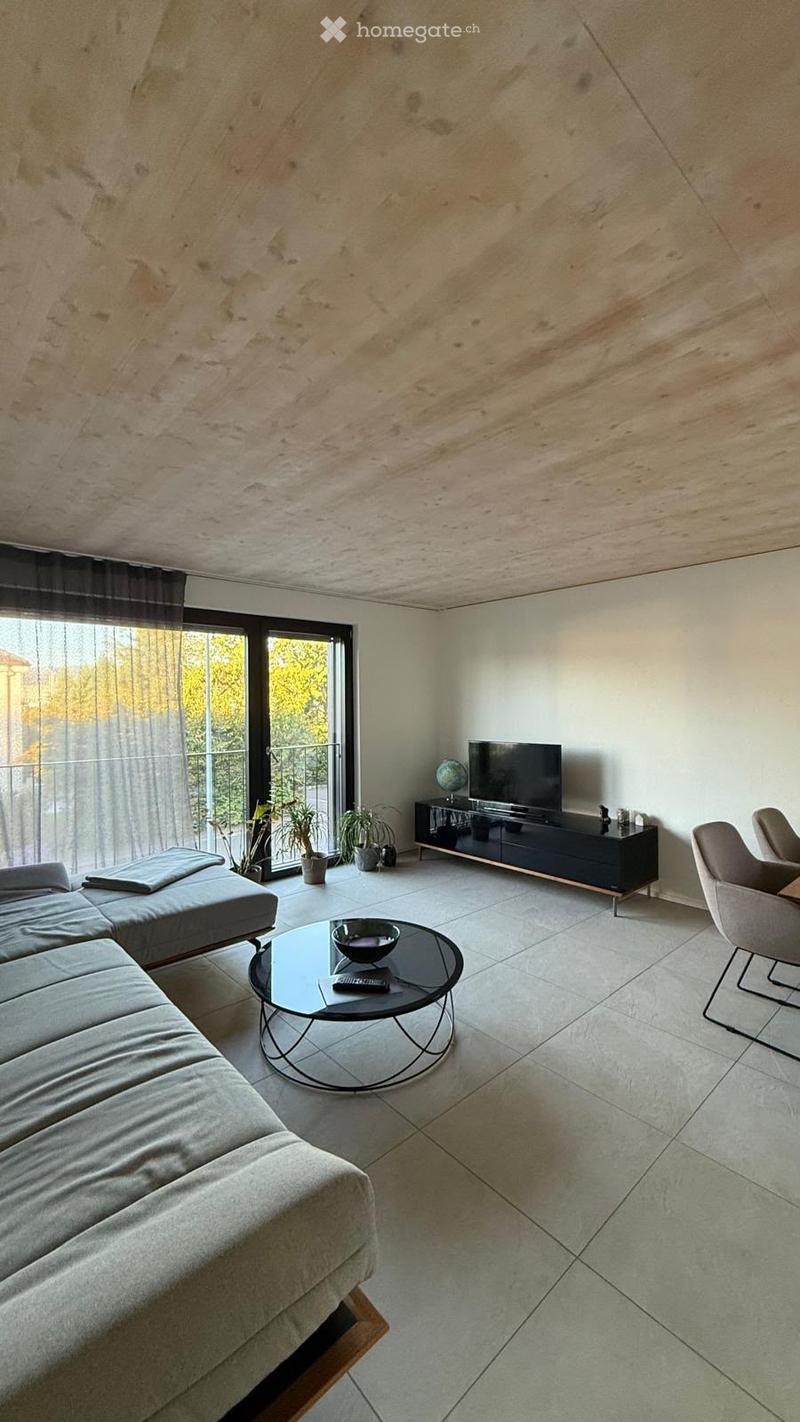 Modern living room with a grey sectional sofa, a round glass coffee table, a TV on a black stand, and a couple of armchairs. The room features large windows with sheer curtains and a view of greenery outside.