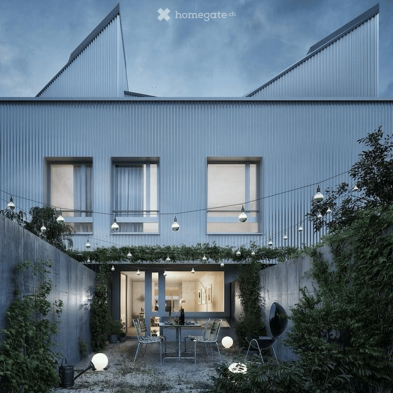 Modern two-story house with grey exterior, large windows, and a small outdoor seating area with a table and chairs surrounded by greenery and string lights.