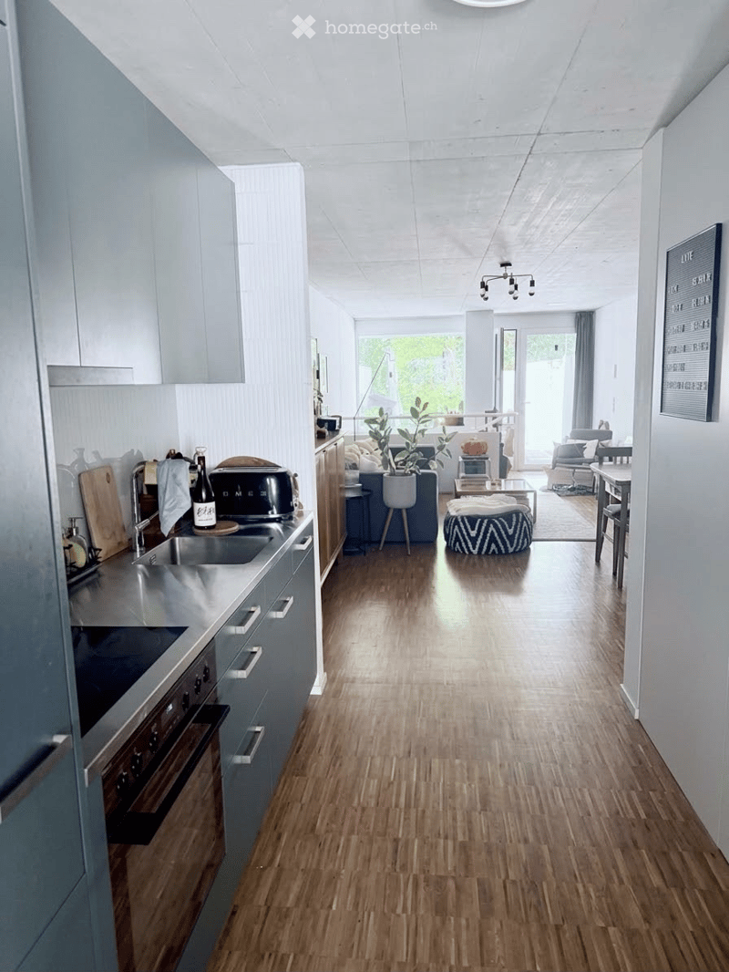 Open-plan kitchen and living area with modern grey cabinets, stainless steel appliances, wooden flooring, and a cozy seating area with a sofa, coffee table, and a patterned ottoman.