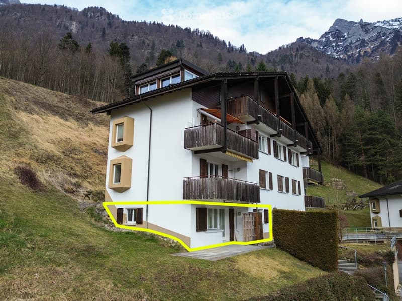 A multi-story white house with wooden balconies and shutters, situated on a hillside with a mountainous backdrop.