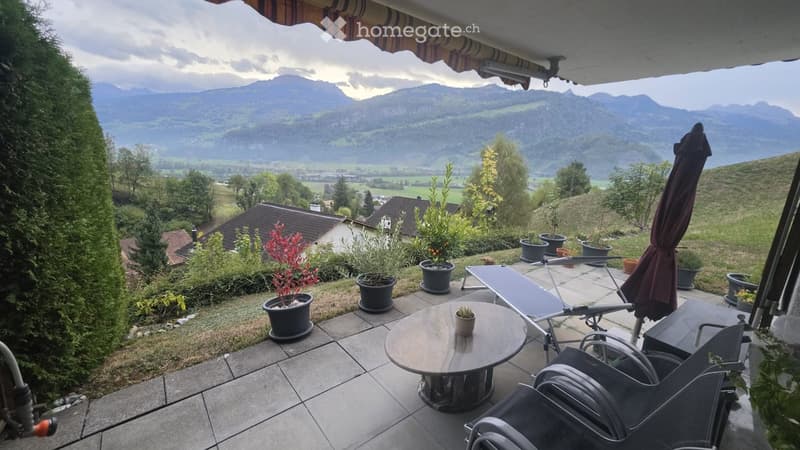 Balcony with a round table, chairs, umbrella, and potted plants, overlooking a scenic mountain view.