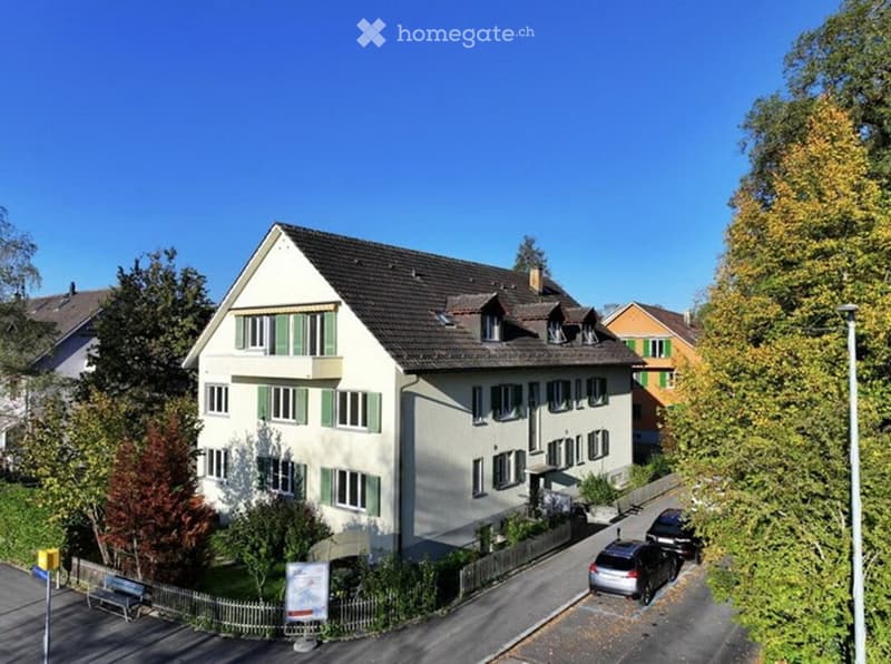 A two-story residential house with a pitched roof, multiple windows, and green shutters. The house is surrounded by trees and has a small garden area with a fence. Several cars are parked on the street in front of the house.