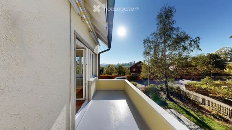 Balcony with white flooring and yellow railing, overlooking a green garden and distant houses under a clear blue sky.