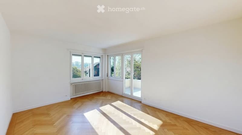 Empty room with wooden flooring, large windows, and a radiator, offering a view of greenery outside.