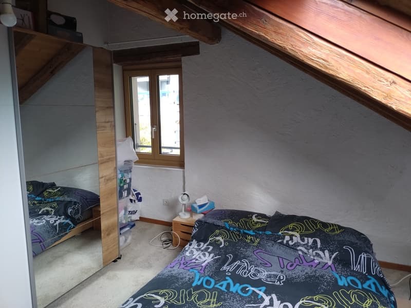 Cozy attic bedroom with slanted wooden ceilings, two single beds with colorful bedding, a nightstand with a lamp, and a shelf with storage boxes.