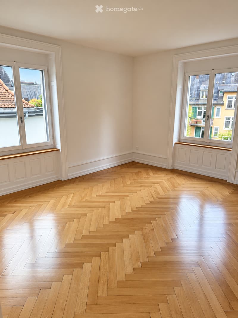 Empty room with light-colored walls, wooden herringbone flooring, and large windows offering a view of the city.