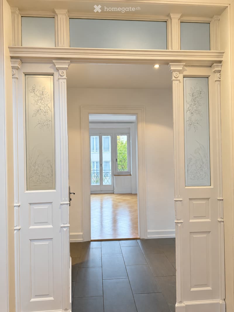 Elegant hallway with white walls, decorative glass panels, polished wooden floor, and a view into a bright room beyond.
