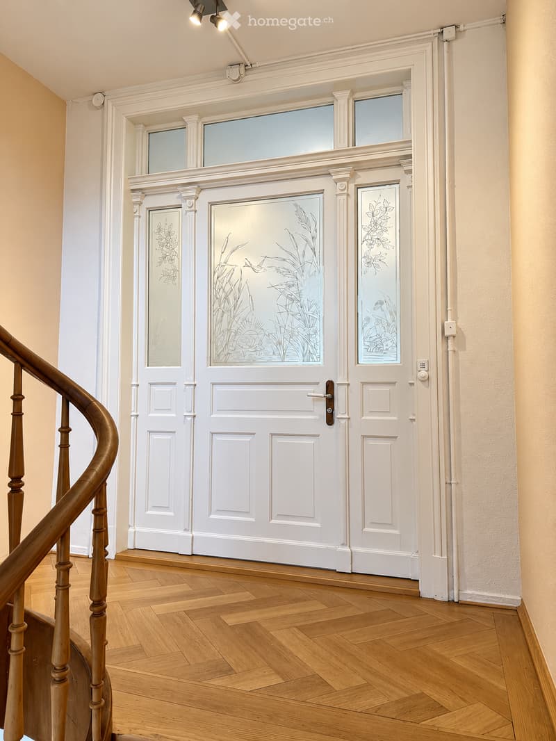 Elegant hallway with wooden flooring, white paneled doors featuring decorative glass panels, and a wooden staircase railing on the left side.
