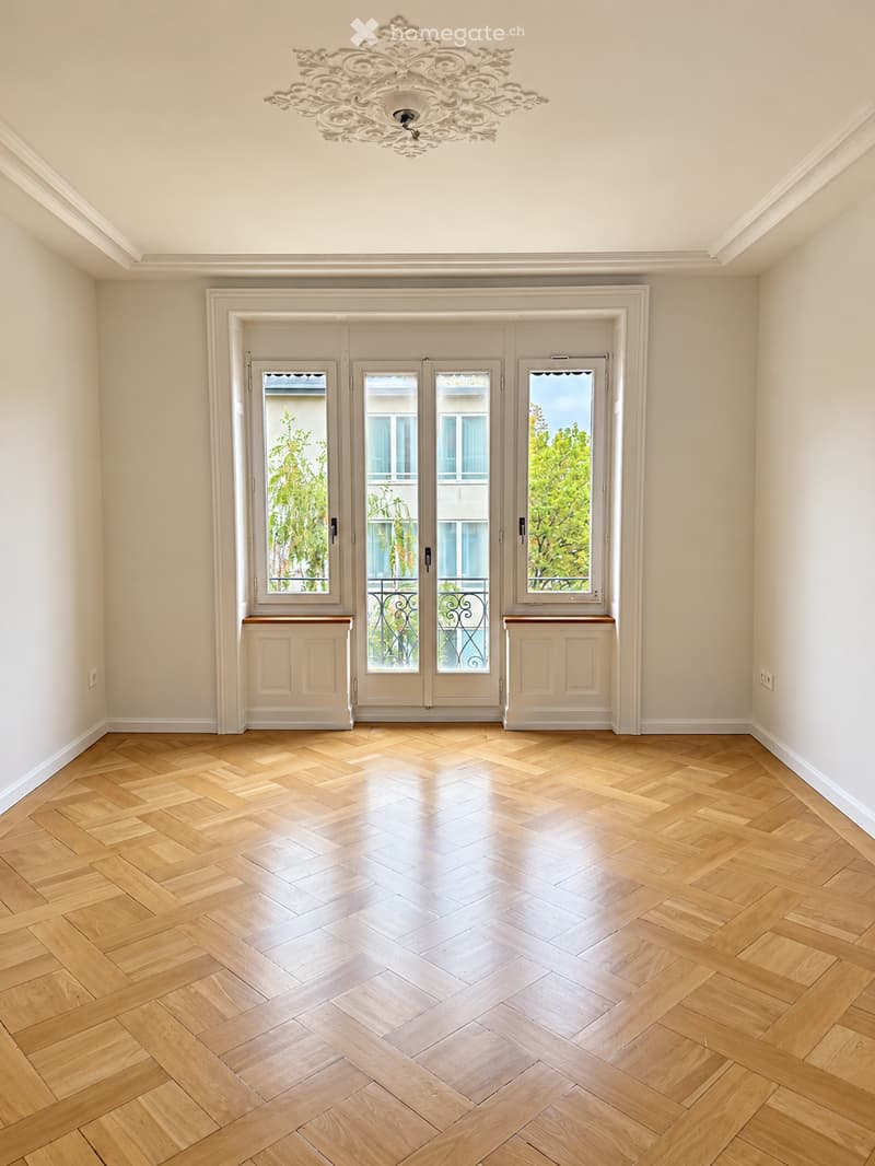 Empty room with light-colored walls, wooden herringbone flooring, and large windows with decorative grilles. A chandelier hangs from the ceiling.