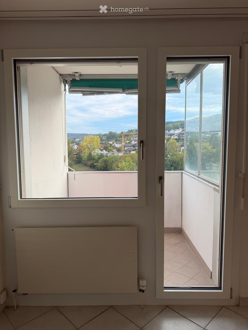 Empty room with tiled floor, radiator, and large windows opening to a balcony with scenic view of town and landscape.