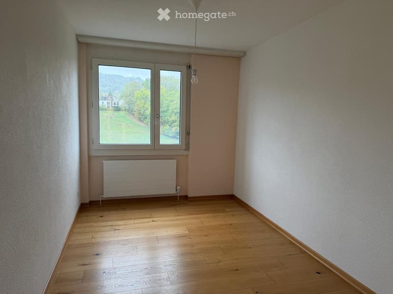 Empty room with wooden flooring, white walls, large window with view, radiator, and a hanging light fixture.