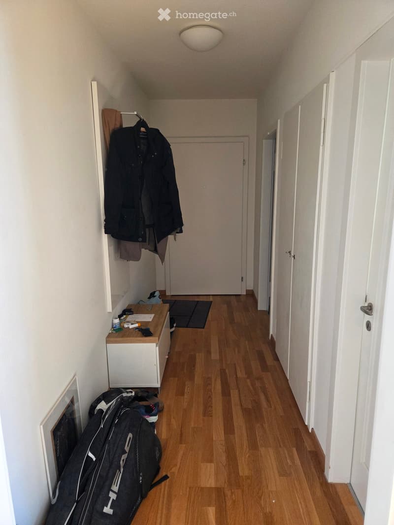 A narrow hallway with wooden flooring, white walls, and several doors. A small wooden bench with various items, including a jacket, a backpack, and some small objects, is placed against the wall. A coat rack with a jacket and a towel is also visible.