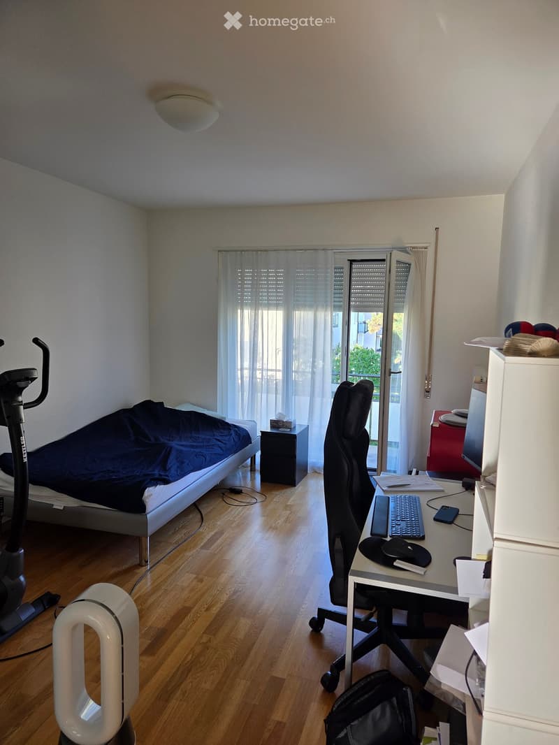 A well-lit bedroom with a bed, desk, chair, and exercise bike. The room features wooden flooring, a large window with white curtains, and a nightstand with a lamp.