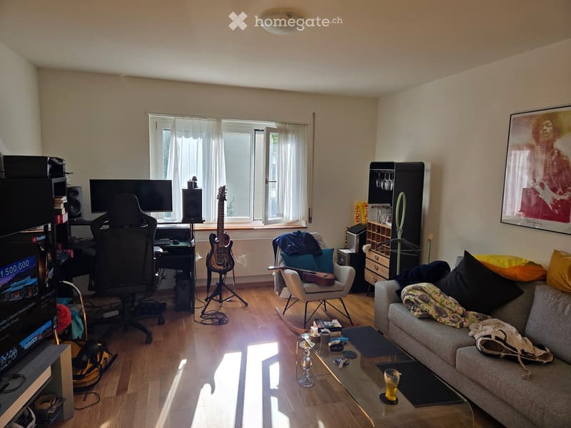 A modern living room with a sofa, coffee table, guitar, and a TV screen. The room features wooden flooring, large windows with white curtains, and a cozy seating area.