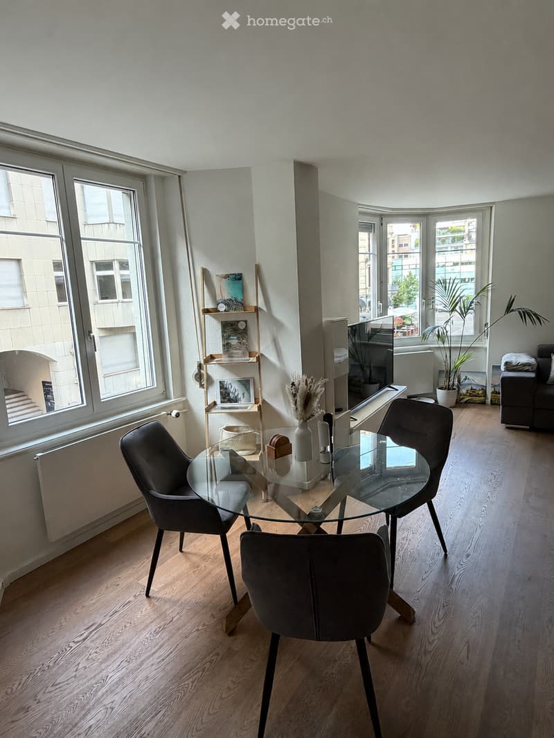 Modern dining area with a glass table, four black chairs, a shelf with decorative items, a vase with dried flowers, and a grey sofa near the window.