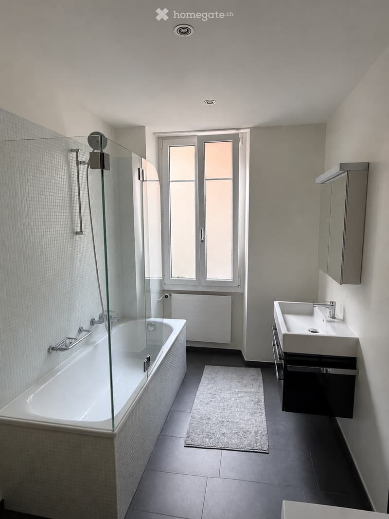 Modern bathroom with a bathtub, glass shower enclosure, sink, mirror, and radiator. Features tiled walls and a large window with frosted glass panes.
