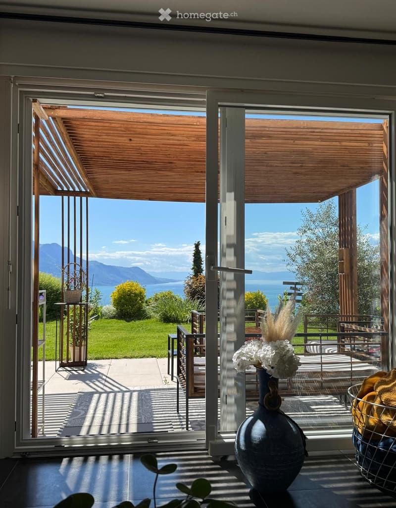 Bright room with large glass doors opening to a patio, featuring a view of a lake and mountains, with a vase and a basket visible inside.
