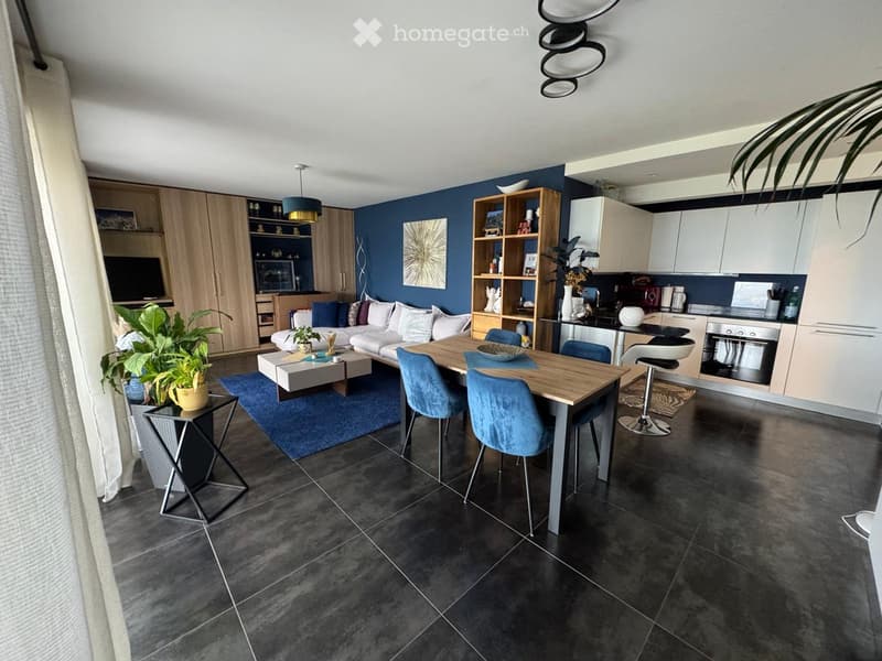 Open-plan living and dining area with a sofa, coffee table, dining table, chairs, and a kitchen area with cabinets, appliances, and a stool.