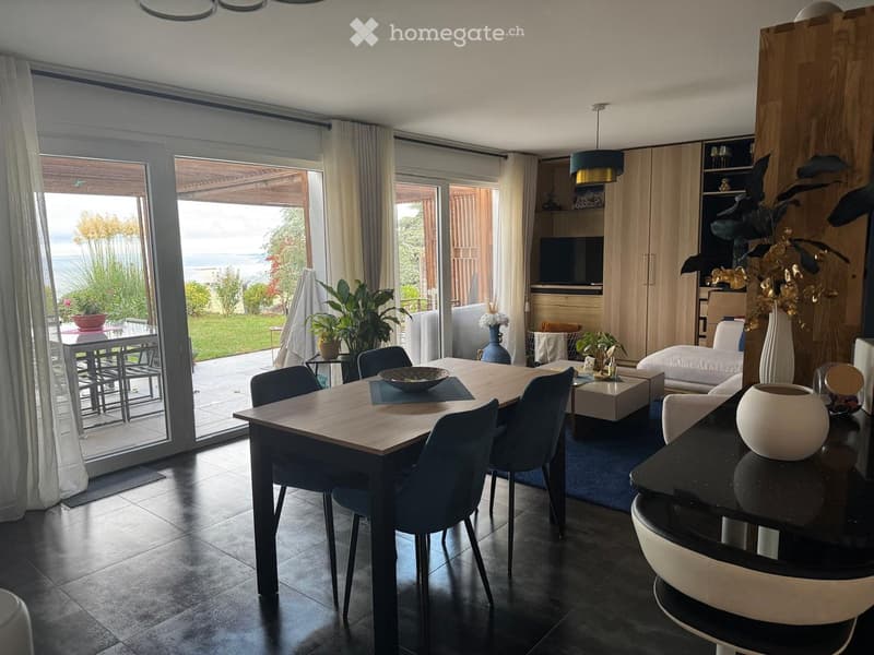 Modern dining and living area with a large wooden dining table, chairs, a sofa, coffee table, and decorative vases. Large windows offer a view of the garden and sea.