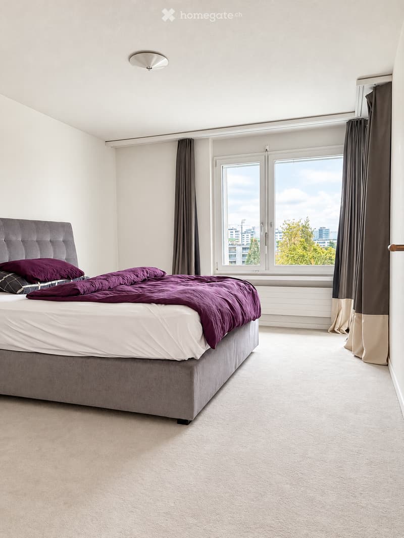 Modern bedroom with a large bed, purple bedding, and large windows with curtains. The room has light-colored walls and carpeted flooring.