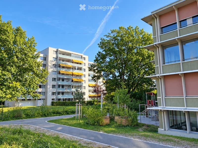 Modern multi-story residential building with balconies and yellow awnings, surrounded by greenery and pathways.