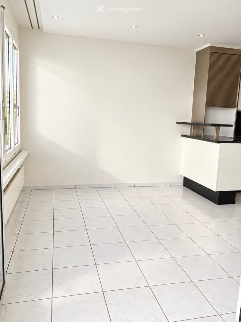 Empty room with white tiled flooring, white walls, and a small built-in cabinet with a countertop. The room has large windows and recessed ceiling lighting.