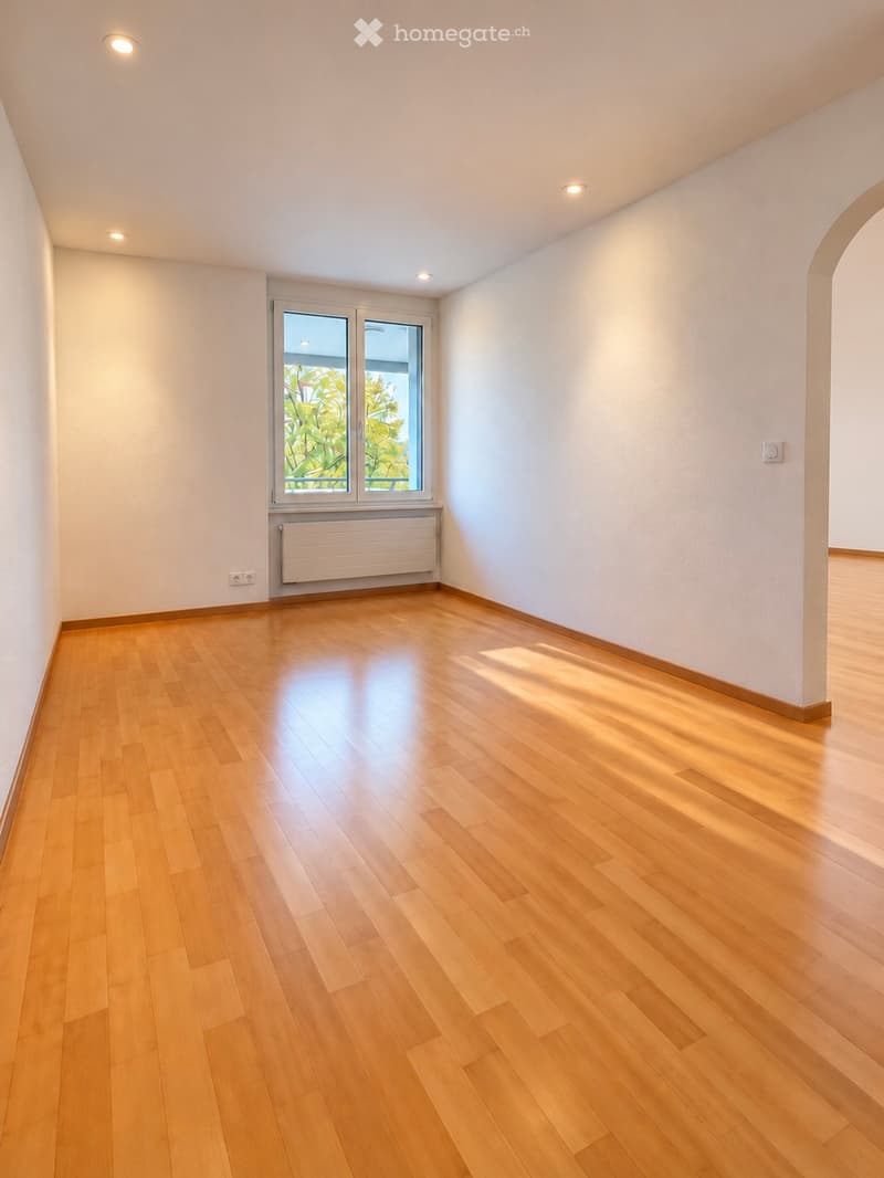 Empty room with light wooden flooring, white walls, and a large window with a view of greenery. The room features recessed ceiling lights and a radiator below the window.