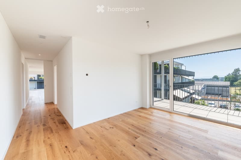 Empty hallway with light wooden flooring, white walls, and large glass doors leading to a balcony with a view of the surrounding area.