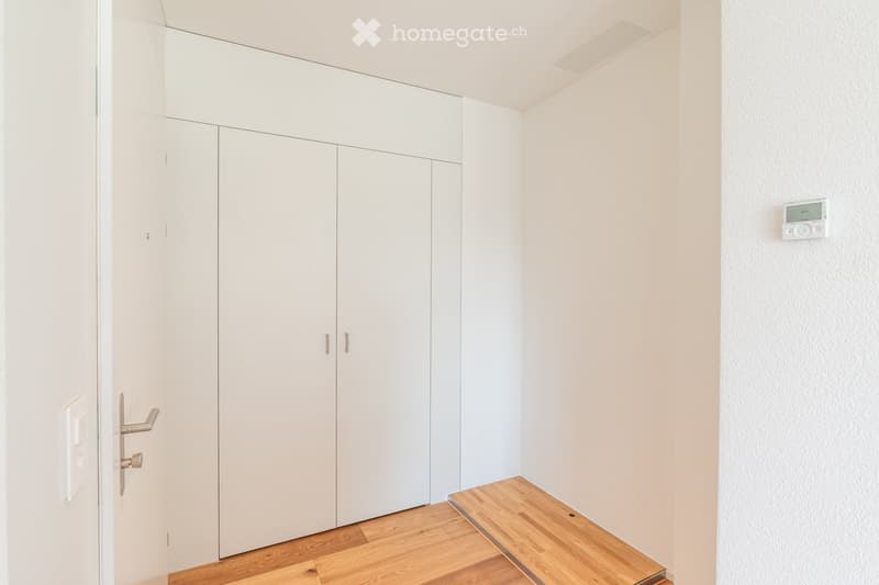 Empty room with white walls, light wooden flooring, built-in white wardrobes, and a small wooden bench near the door.