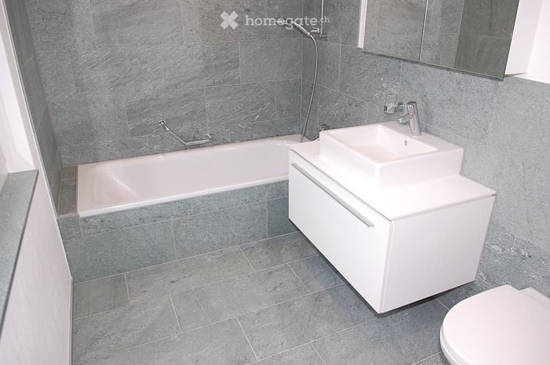 Modern bathroom with grey tiled walls and floor, featuring a bathtub, a white sink with a cabinet, a toilet, and a mirror above the sink.