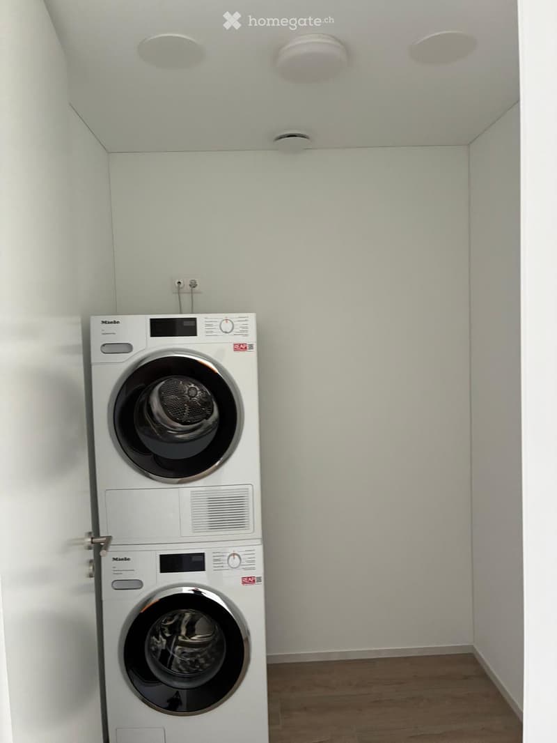 A small, well-lit room with a stacked washing machine and dryer unit against a white wall, wooden flooring, and recessed ceiling lights.
