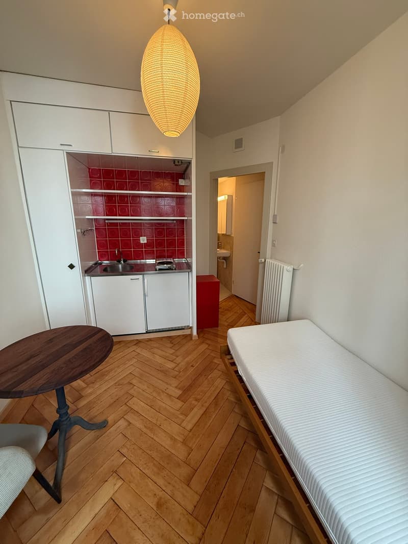 A compact studio apartment with a bed, small table, chair, and a kitchenette area featuring white cabinets, a sink, and a faucet. The floor is wooden, and a radiator is visible near the door.