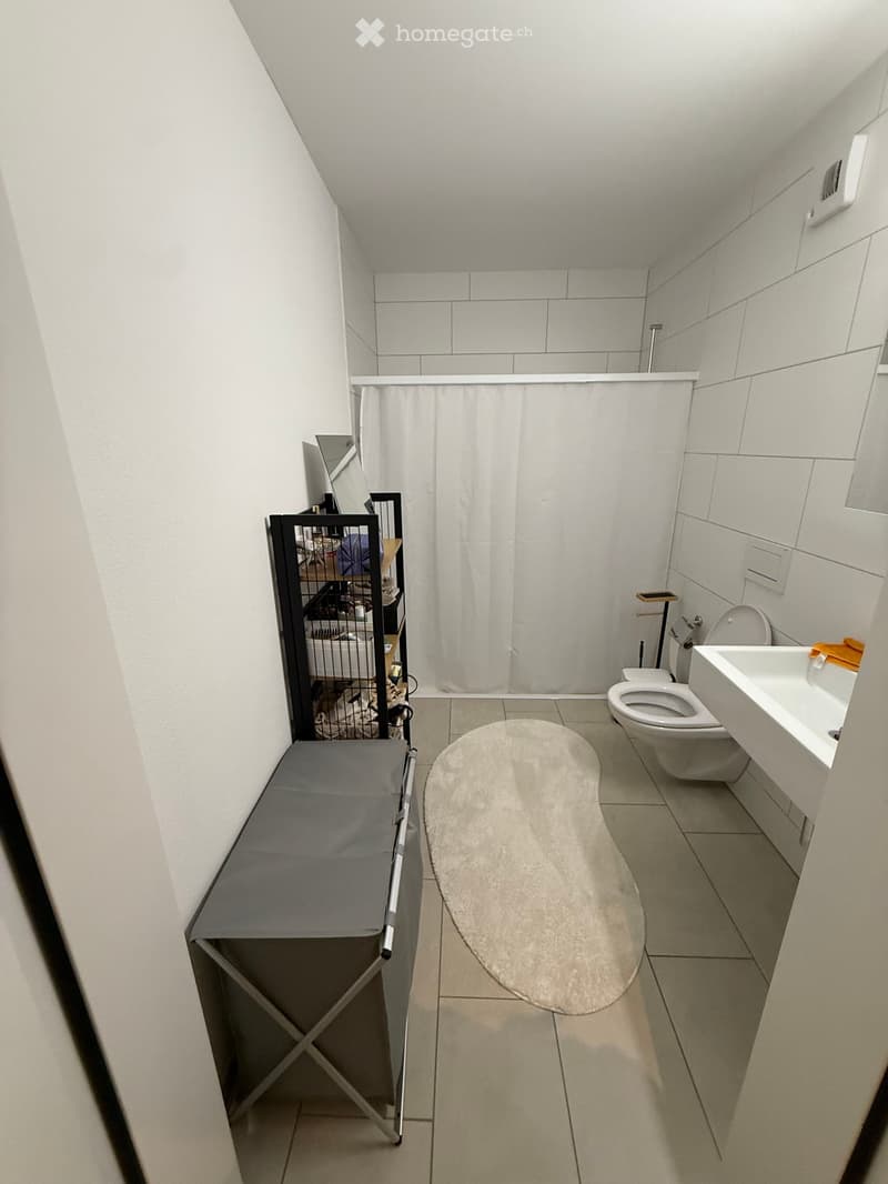 Modern bathroom with white tiles, a toilet, sink, mirror, shower curtain, bath mat, and a small cage with a table nearby.