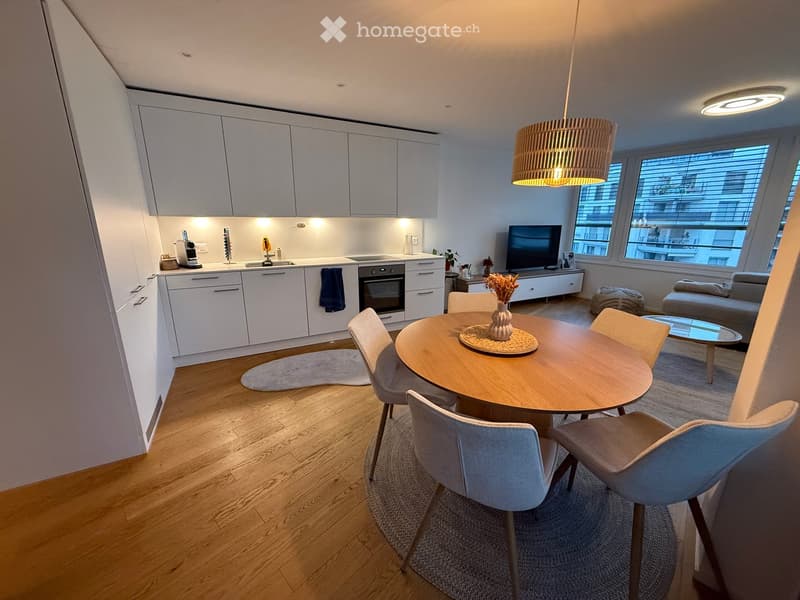 Modern, open-plan living and dining area with a round wooden dining table, four chairs, a sofa, a coffee table, and a kitchen with white cabinets, an oven, and a refrigerator. The space features wooden flooring, pendant lighting, and large windows with bl