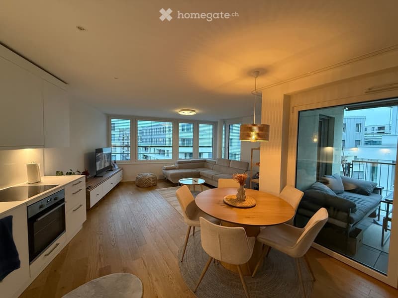 Modern, open-concept living and dining area with a sofa, coffee table, dining table with chairs, and a small kitchen area with an oven and cabinetry.