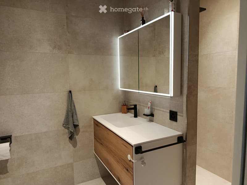 Modern bathroom with a white countertop sink, wooden cabinet, illuminated mirror, and a towel hanging on the wall.