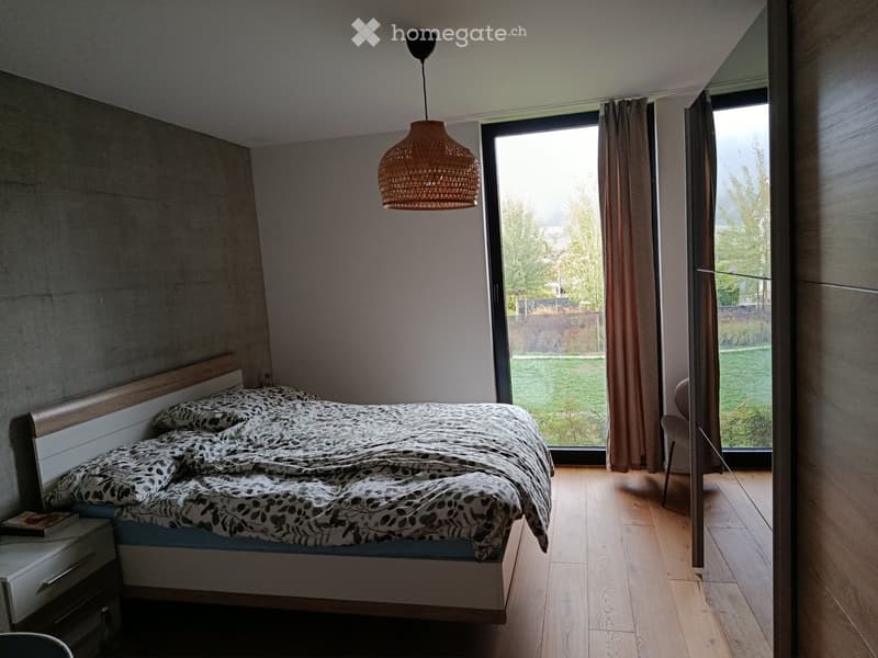 Modern bedroom with wooden flooring, large window with view, bed with patterned bedding, nightstand, chair, and pendant light.