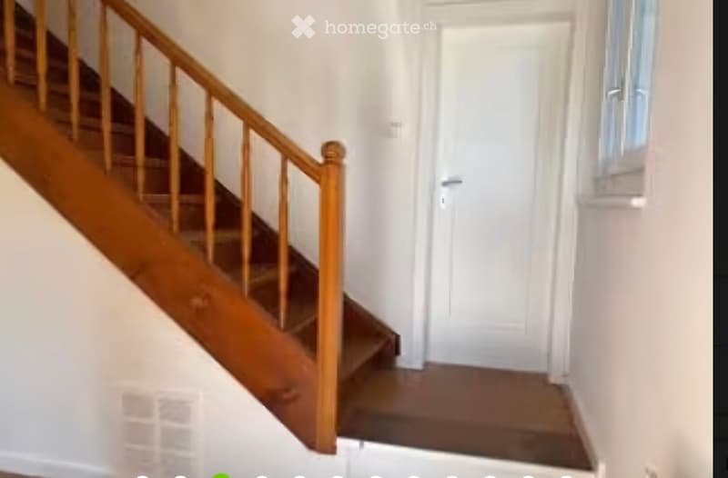 A narrow hallway with a wooden staircase leading upwards, featuring a wooden handrail and a white door at the end.